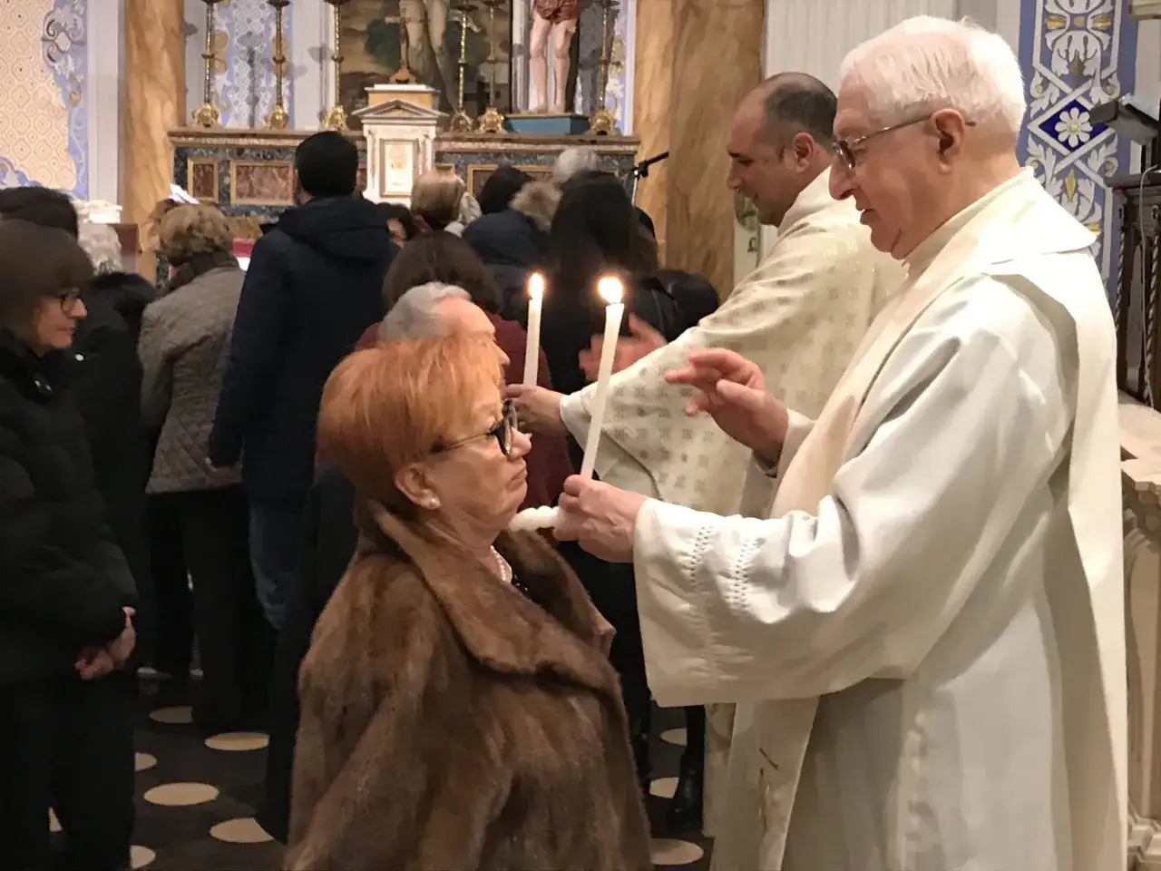 Messa in chiesa con candele accese. Un sacerdote benedice una donna con una candela, invocando il santo protettore della gola.