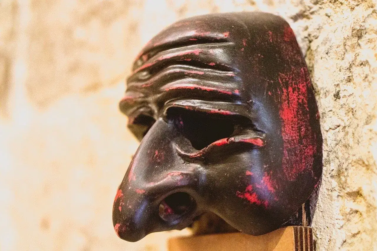 Maschera di Pulcinella, simbolo del carnevale, con tratti scuri e rossi consumati, appoggiata su uno sfondo chiaro e ruvido.