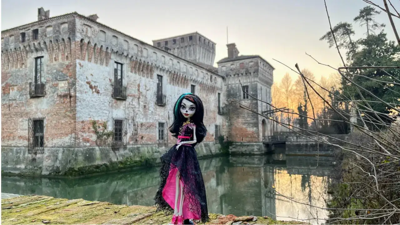 Una bambola con un vestito nero e rosa, simile a una dama bianca, posa davanti a un antico castello con un fossato.