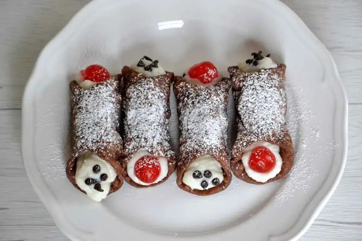 Cannoli ricetta: quattro dolci siciliani al cioccolato, ripieni di crema e decorati con ciliegie candite e gocce di cioccolato.
