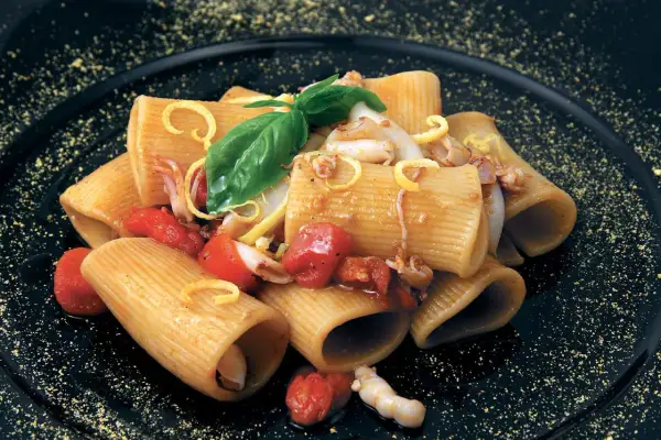 Rigatoni con calamari e pomodorini pachino, guarniti con basilico e scorza di limone su piatto nero.