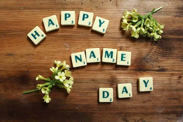 Tessere del gioco compongono "HAPPY NAME DAY" su un tavolo di legno, con fiori gialli che adornano la scena.