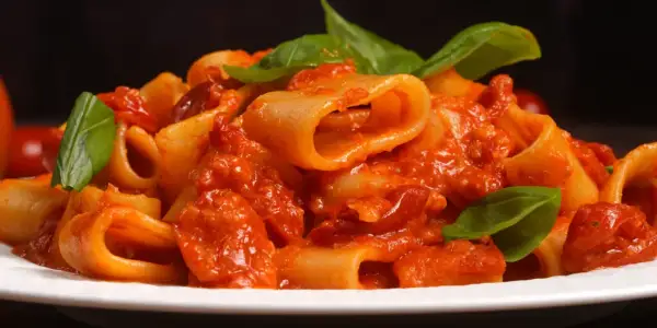 Un piatto di rigatoni allo scarpariello, conditi con un sugo rosso ricco e guarniti con foglie di basilico fresco.