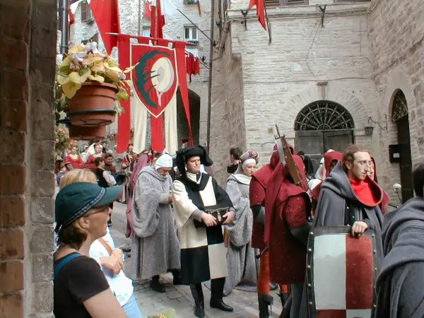 Persone in costume medievale sfilano in una strada acciottolata, con stendardi rossi e bianchi che sventolano. La scena evoca il **calendimaggio significato**, una festa di primavera.