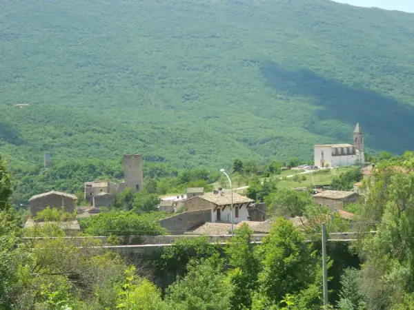Il gigante di acciaio veglia su un borgo antico, tra torri e case di pietra, sotto un monte verdeggiante.