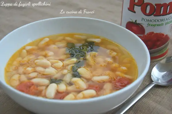 Zuppa di fagioli spollichini fumante in una ciotola bianca, con pomodori e verdure. Accanto, una lattina di pomodori e un cucchiaio.