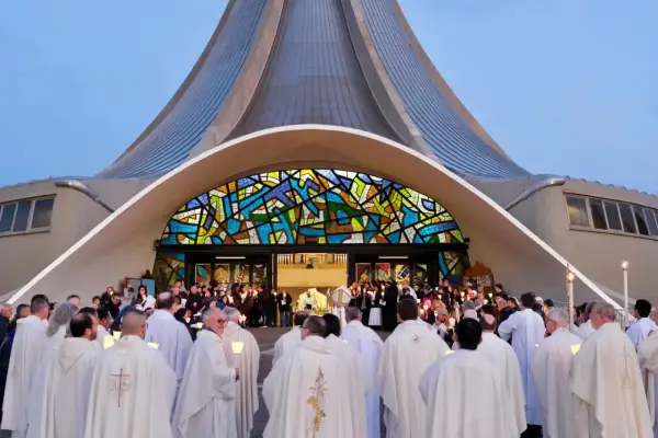 Un gruppo di sacerdoti, con candele accese, si riunisce davanti a una chiesa moderna con una vetrata colorata. La luce delle candele illumina i loro volti, quasi a riflettere il volto di Gesù.